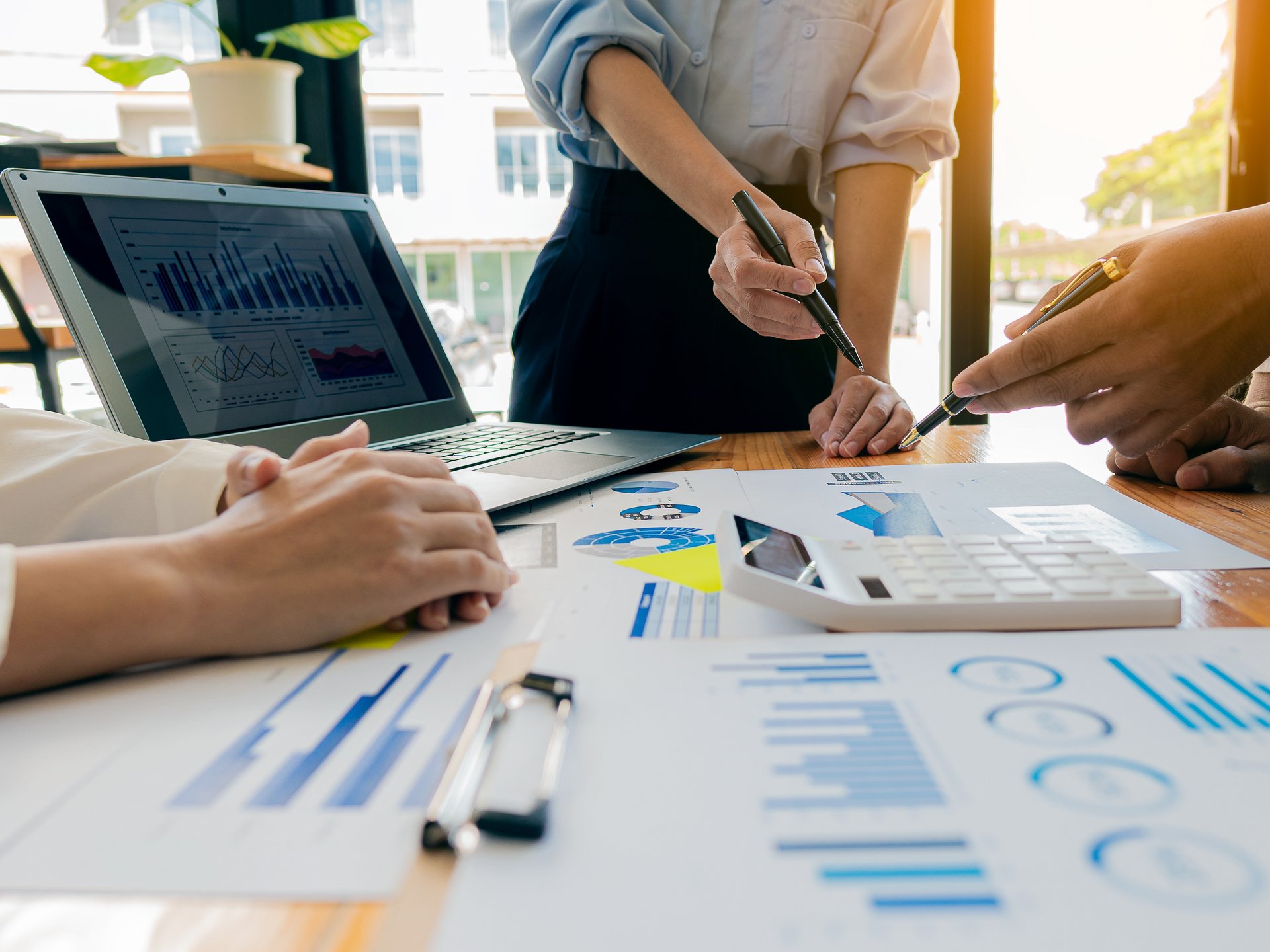meeting situation: in the foreground a table with docuemnts and charts, a laptop and two hands that point towards a chart in the background