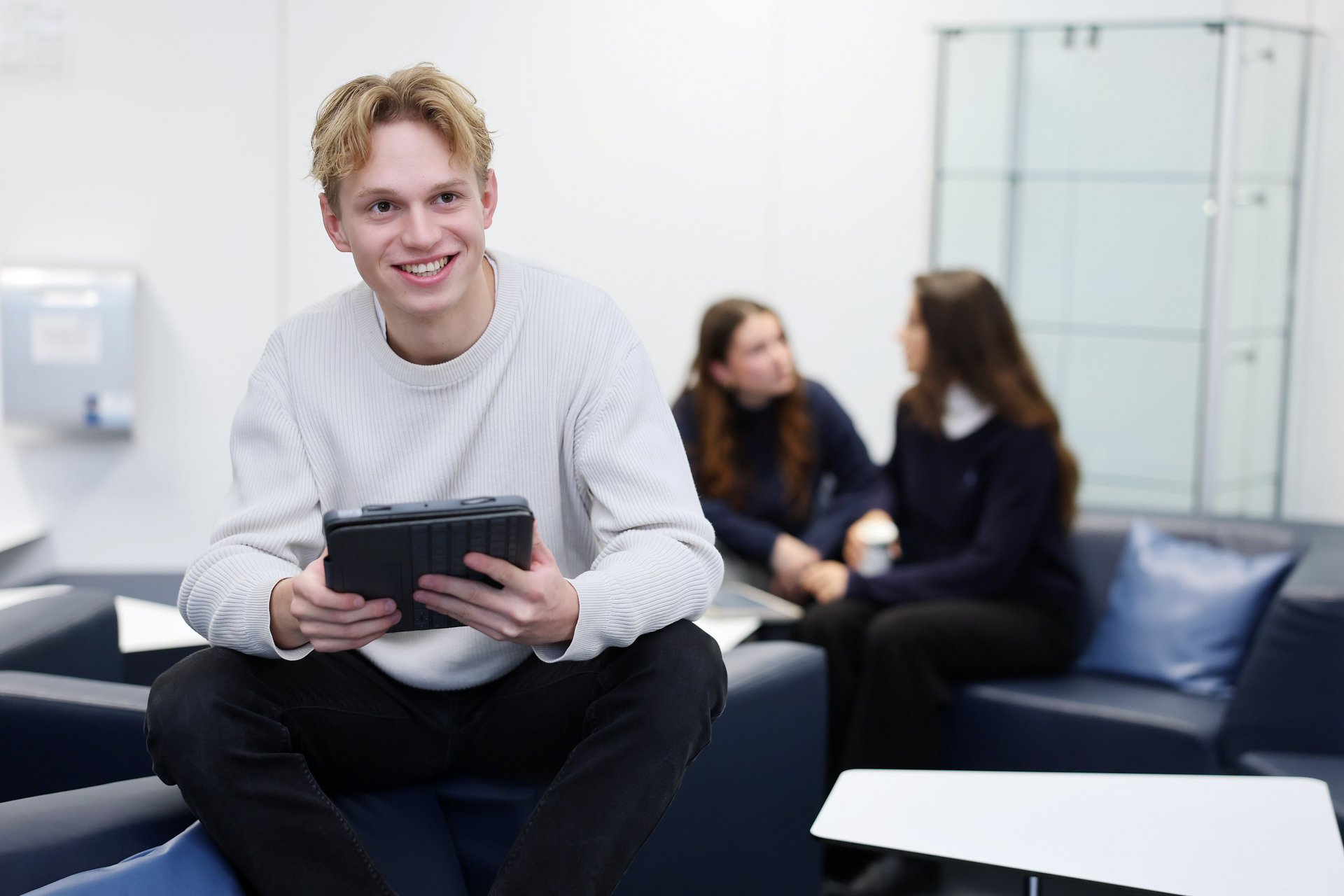 Student sitzt mit Tablet, im Hintrgrund sprechen 2 Frauen